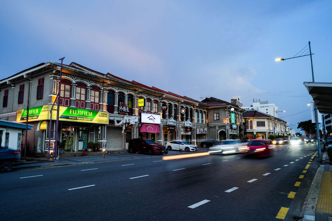 A street of shophouses at dusk, with traffic passing
